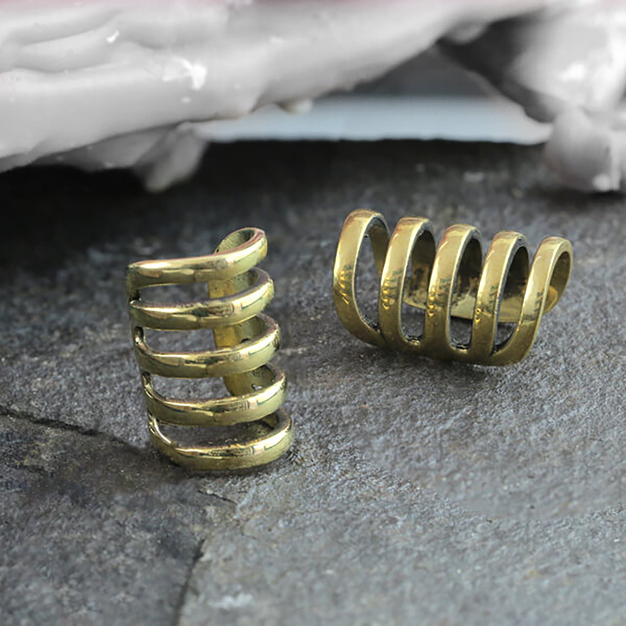 Two KIRA Long Cage Ear Cuffs in Gold by Kaya Movement are displayed on a textured stone surface. One ear cuff stands upright while the other lies flat, showcasing their unique, minimalist design. The adjustable-size cuffs evoke the elegance of finely crafted accessories. In the background, part of a larger object or decoration is partially visible.