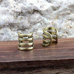 Two KIRA Small Cage Ear Cuffs in Gold by Kaya Movement, each featuring a minimalist design, are displayed on a wooden surface against a textured stone background. The ear cuff on the left has a horizontal spiral design, while the one on the right showcases an angled, diagonal pattern. Each ear cuff boasts an adjustable size for a perfect fit.