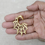 A person holds a small, gold-colored metal hook shaped like a spider web. The intricate design of the web spans from the hook, showcasing its detailed and artistic craftsmanship. These resemble the Spider Web Gothic Steel Ear Hangers in Gold | 2 gauge by Kaya Movement. The background is a neutral grayish surface.