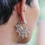 A close-up of an ear wearing the Spider Web Gothic Steel Ear Hangers in Silver from Kaya Movement, designed to resemble spider web ear weights. The intricate web-like structure of the 2 gauge earring spreads outward in a semi-circular shape. The background is blurred, focusing attention on these unique ear weights.