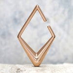 The Minimalist Diamond Ear Hangers in Rose Gold by Kaya Movement are displayed upright on a white textured surface against a blurred neutral background. The hangers feature a unique angular design with sharp edges and an open gap on one side, reminiscent of tribal ear weights.