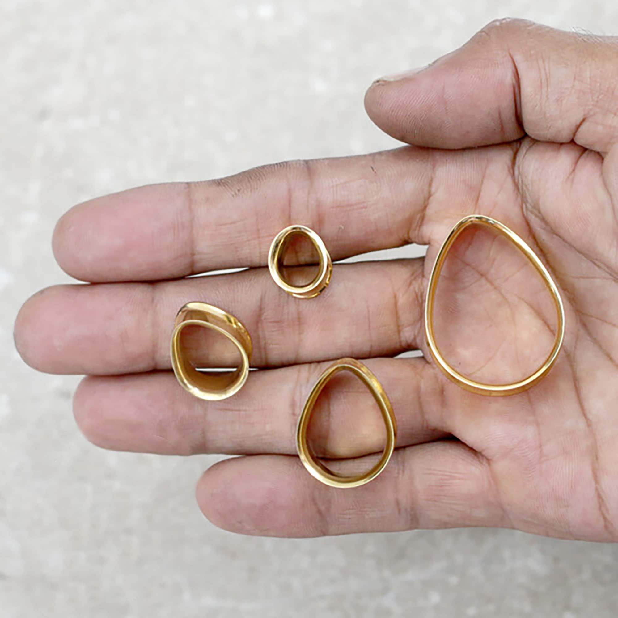 A hand holds four golden, teardrop-shaped plugs of various sizes against a light-colored, textured background. The largest plug is near the thumb, and the smallest is near the tip of the middle finger. All plugs feature smooth, polished surfaces, reminiscent of surgical steel used in tribal plug designs from Kaya Movement's Teardrop Tunnel collection, which includes a range from 3/4 gauge tunnels to 00 gauges.