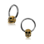 Two Kaya Movement SKULL Gothic Silver Rings with Gold Pendants are displayed against a white background. Each ring, available in 18, 16, or 14 gauge and crafted from surgical steel, features a small, intricately detailed skull with hollow eye sockets, a nose cavity, and teeth detailing. The design adds a gothic or edgy aesthetic to these striking pieces of body jewelry.