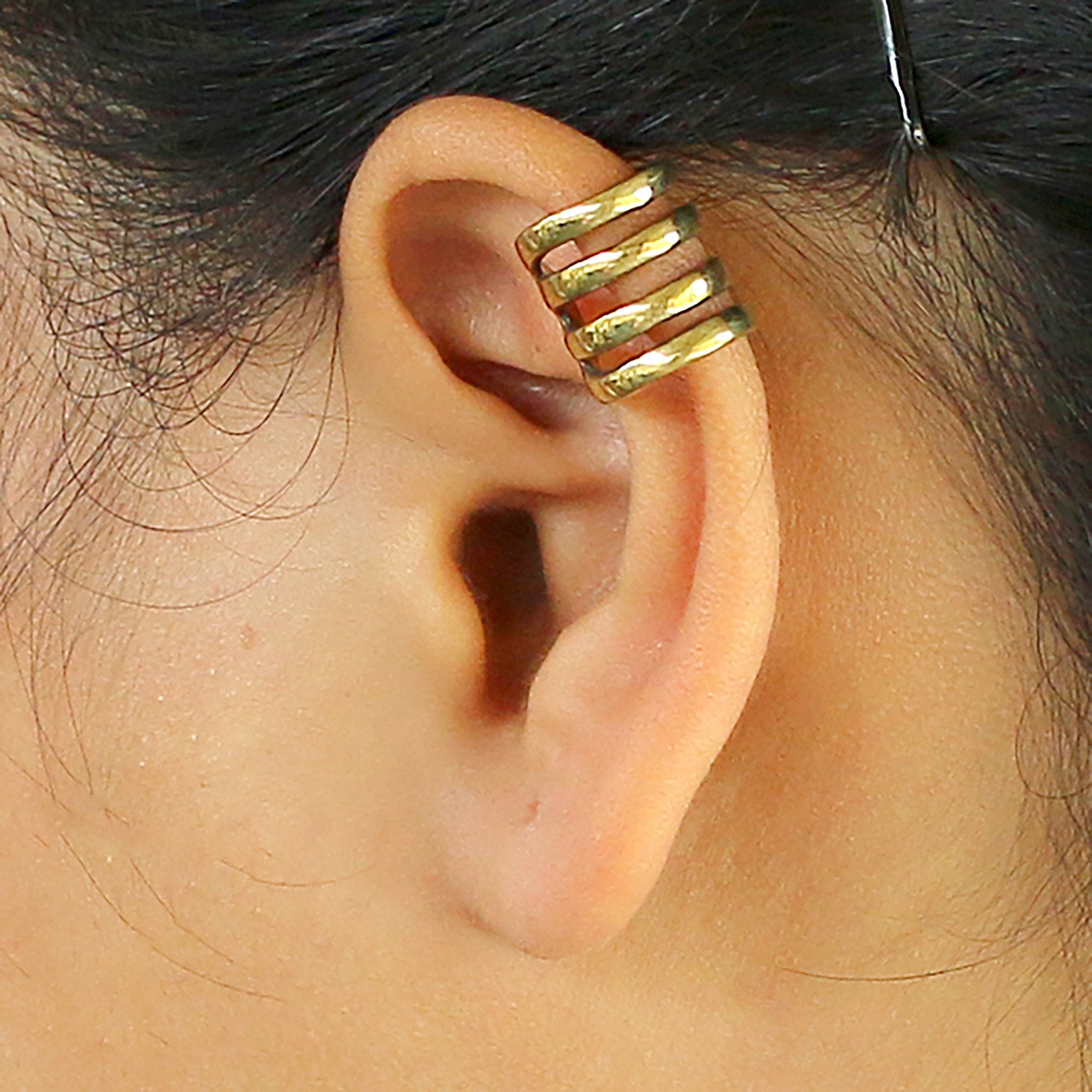 Close-up of a person's ear featuring the KIRA Small Cage Ear Cuff in Gold by Kaya Movement, worn on the upper part of the ear. The minimalist design boasts a smooth finish and hugs the ear without needing a piercing. With an adjustable size for a perfect fit, the person's hair is partially visible.