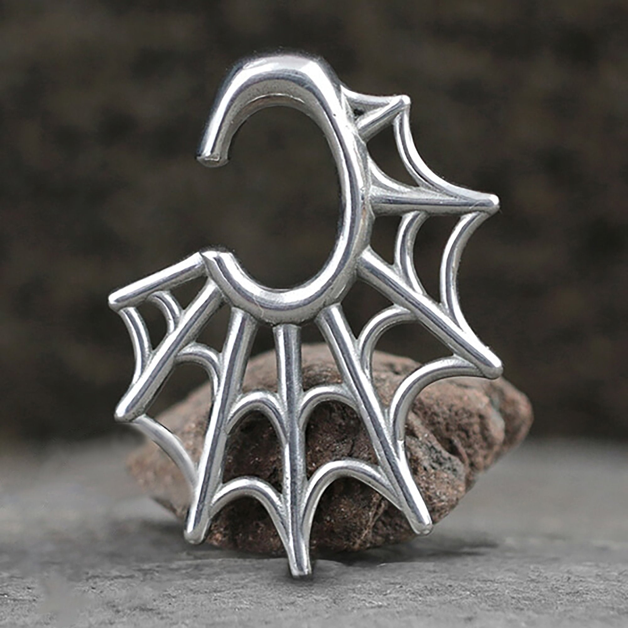 The Spider Web Gothic Steel Ear Hangers in Silver | 2 gauge by Kaya Movement, designed to resemble an intricate spider web with web-like details extending from the main circular part, are displayed against a neutral, rocky background. Crafted from surgical steel ear weights, these hangers present a modern and gothic style.