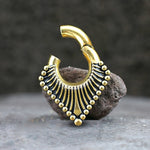 A gold-plated Steel Triangle Ear Weight by Kaya Movement, adorned with intricate black detailing, sits sculpturally on a gray rock. The 2-gauge ear weight features a semi-circular design with descending gold beads and lines fanning out from the center, creating an elegant and artistic aesthetic reminiscent of traditional tribal ear weights.