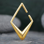A sleek, geometric gold-plated PVD pendant from the Kaya Movement, resembling an angular, stylized letter "G" or an arrowhead, rests on a smooth gray stone background. The 2 gauge Minimalist Diamond Ear Hanger has sharp, clean lines and a polished finish that stand out against the muted backdrop.