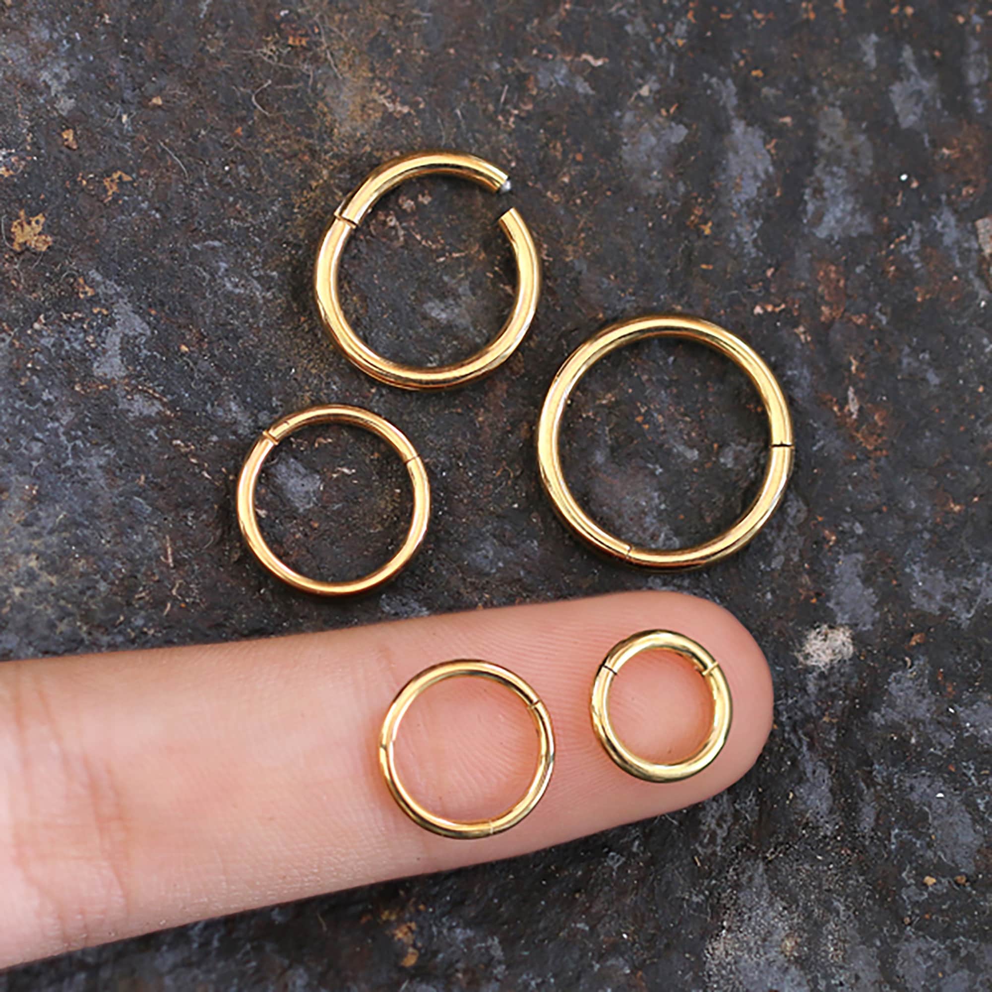 A finger pointing at five AMOI Clicker Nose Rings in Gold by Kaya Movement of varying sizes placed on a textured dark surface. The middle finger bearing two gold hoops illustrates the size difference, with three more hoops arranged above the finger.