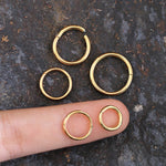 A finger pointing at five AMOI Clicker Nose Rings in Gold by Kaya Movement of varying sizes placed on a textured dark surface. The middle finger bearing two gold hoops illustrates the size difference, with three more hoops arranged above the finger.