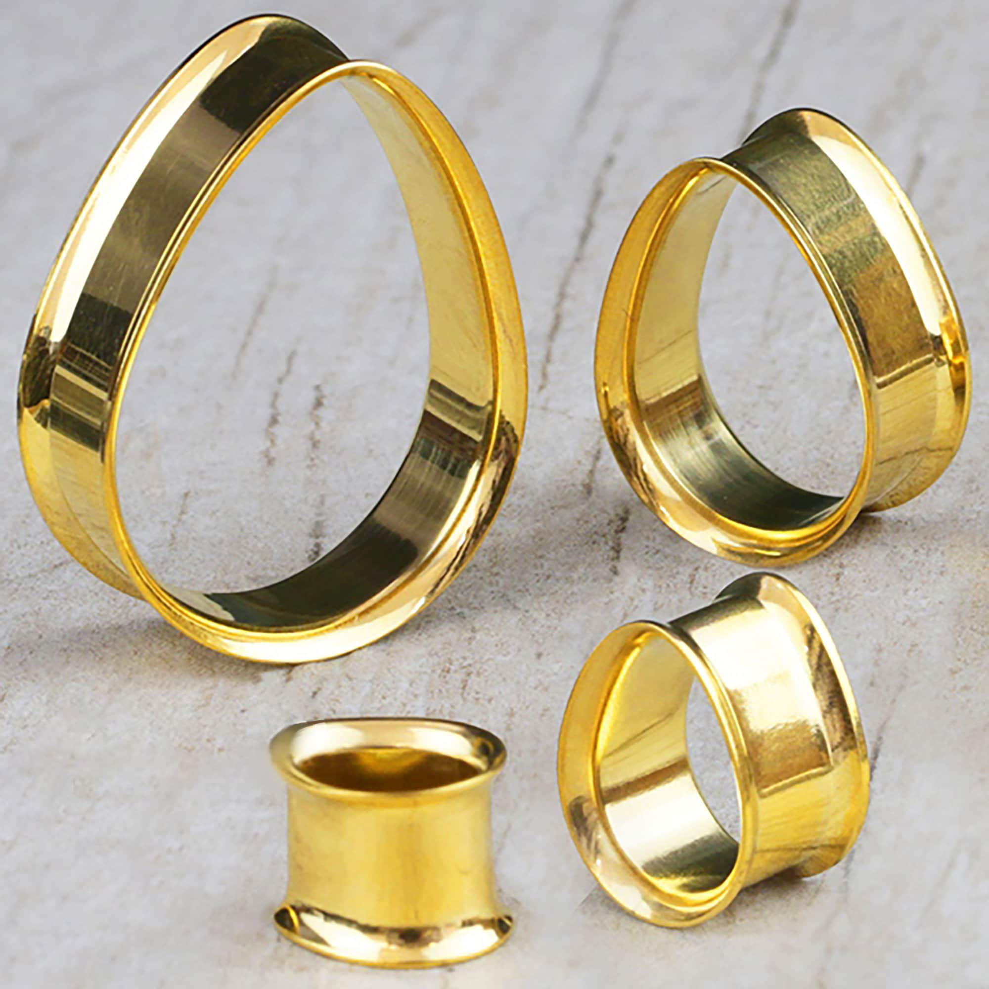 A close-up of four Teardrop tunnels from Kaya Movement made of shiny gold metal, reminiscent of a tribal plug, arranged on a light gray textured surface. The plugs vary in size, showcasing their unique twisted design that creates a single continuous surface.