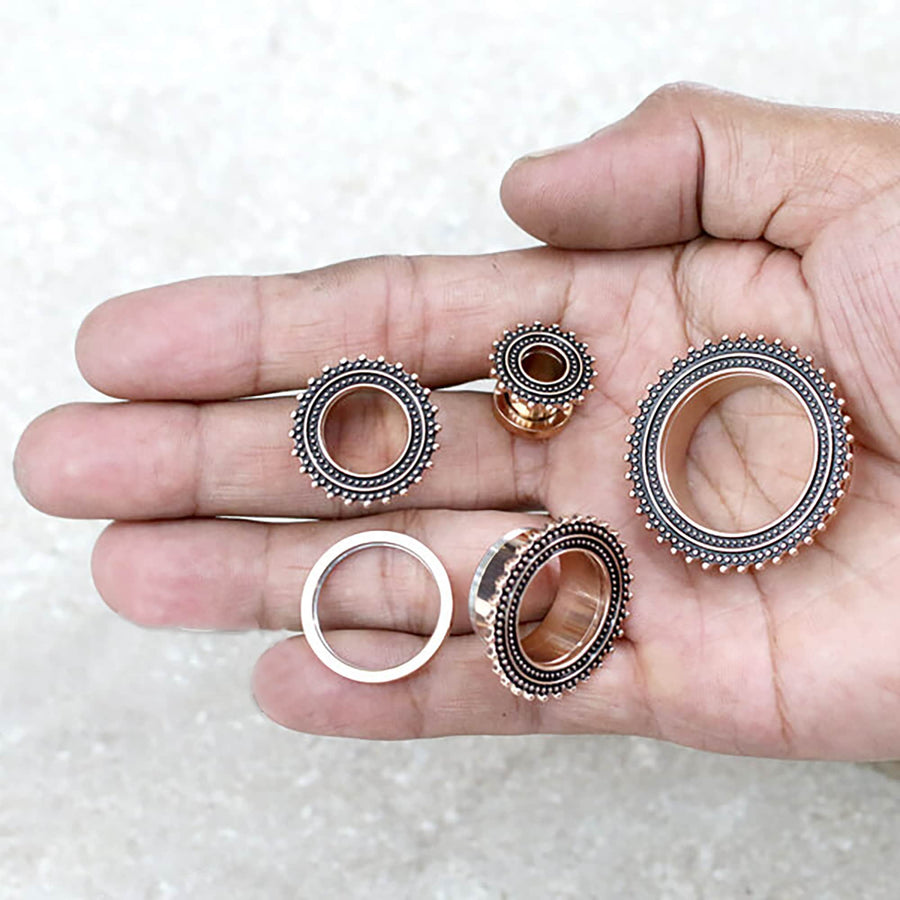 A hand holding three intricate steampunk-style gears and two Kaya Movement Rose Gold Plugs, designed as Flesh Tunnel piercings with beaded detailing, reminiscent of tribal plugs. The metallic rose gold finish showcases detailed craftsmanship against an off-white textured surface.