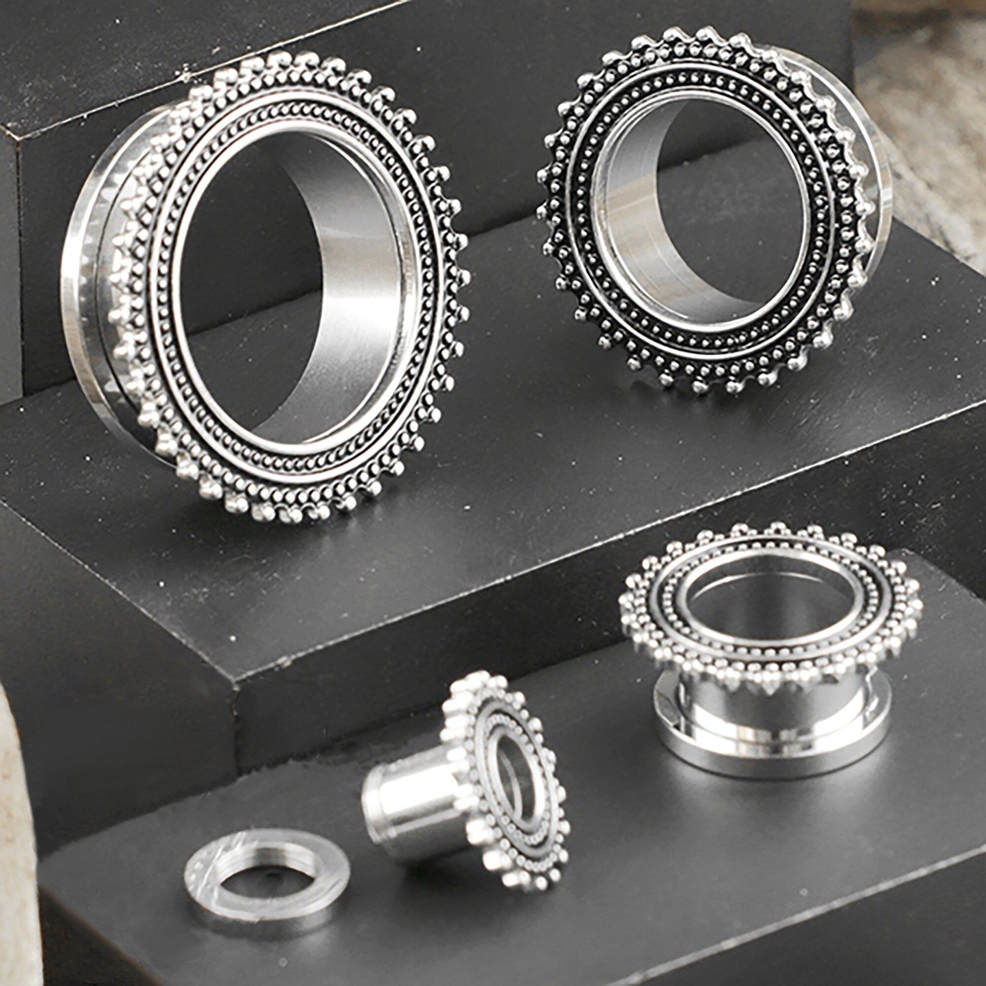 A photograph displaying four metallic, circular items, each detailed with a decorative, beaded edge. These items are ear gauges from Kaya Movement, specifically stainless steel plugs in silver. They appear to be 2g (6mm) to 00g gauges and could also be 8 gauge screw tunnel pieces intended for stretched ear piercings. They are neatly arranged on a layered, black surface.