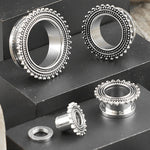 A photograph displaying four metallic, circular items, each detailed with a decorative, beaded edge. These items are ear gauges from Kaya Movement, specifically stainless steel plugs in silver. They appear to be 2g (6mm) to 00g gauges and could also be 8 gauge screw tunnel pieces intended for stretched ear piercings. They are neatly arranged on a layered, black surface.