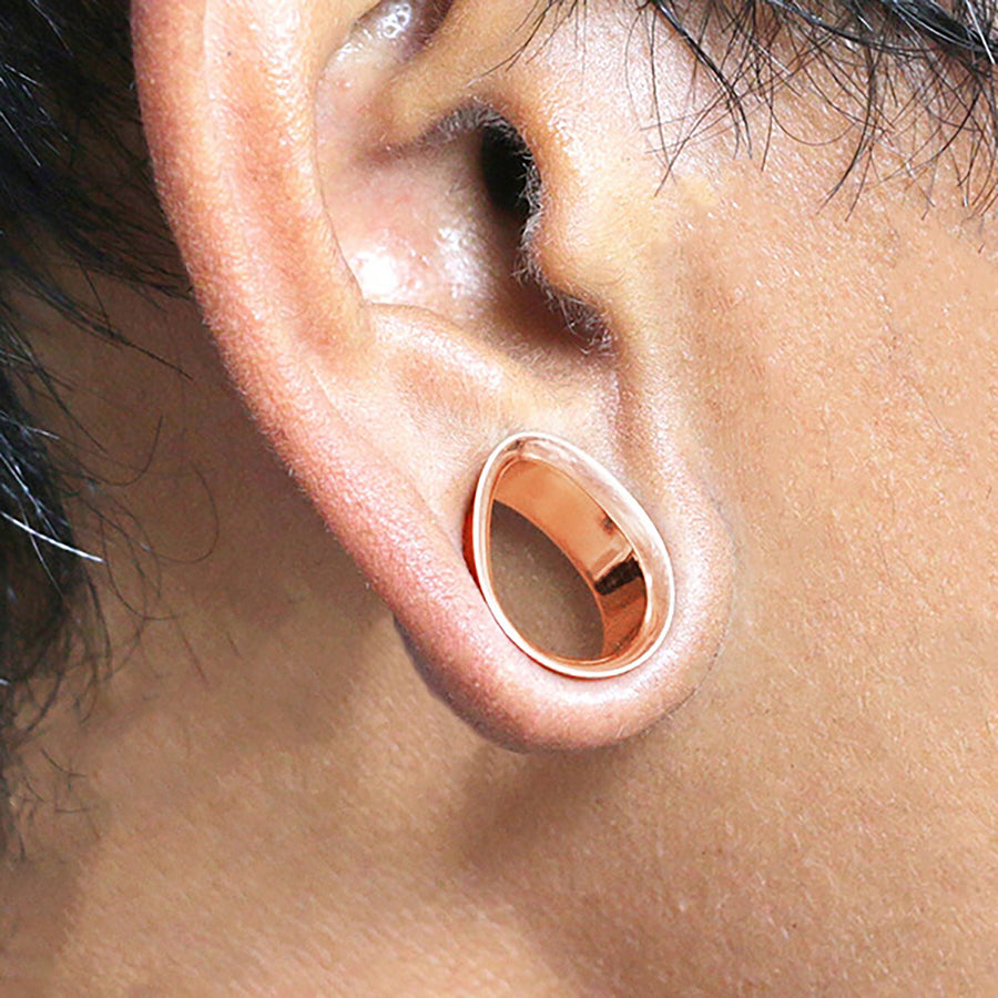 A close-up shot of a person's ear adorned with a Kaya Movement Teardrop Tunnel Plug in rose gold, inserted into the earlobe as a 0 gauge piercing. The hollow center of the flesh tunnel reveals the skin within, while the surrounding area appears slightly reddish. Strands of hair are partially visible around the ear.