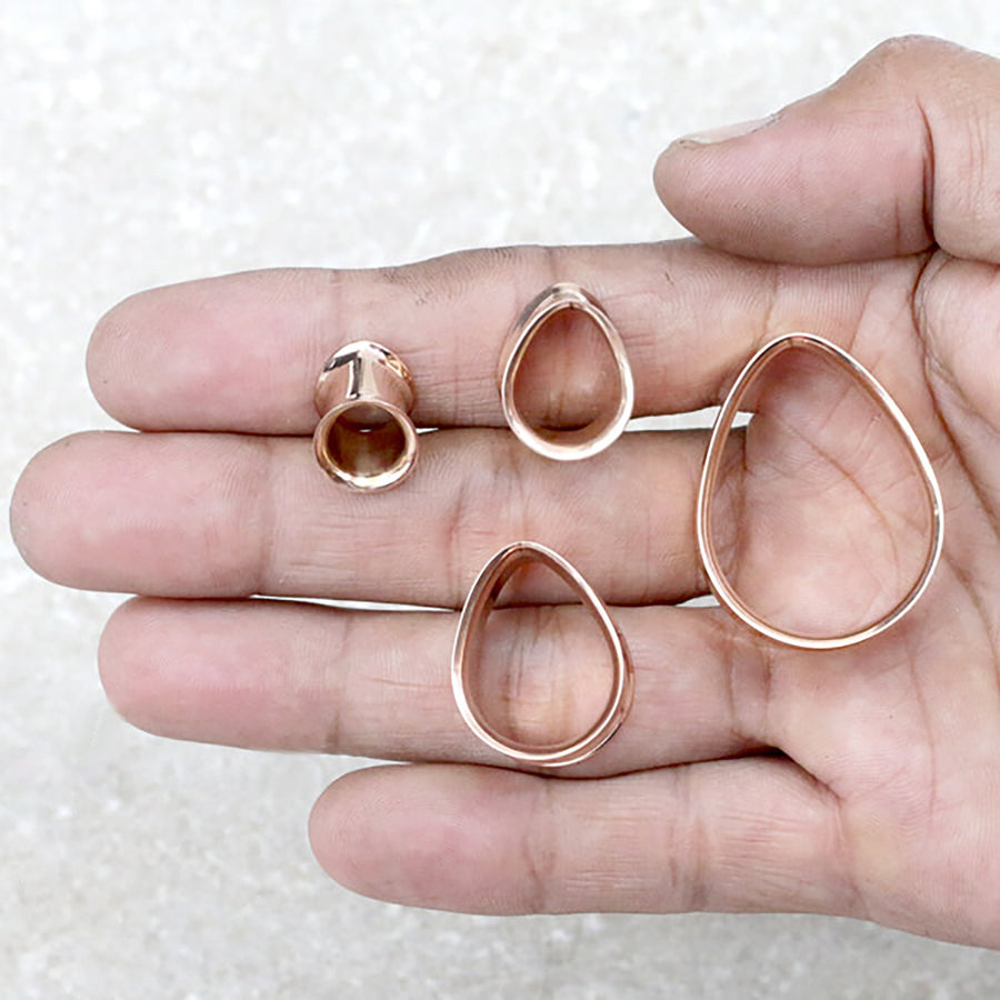 A hand is holding four rose gold metallic shapes resembling components of handcrafted tribal plugs by Kaya Movement: a small strip, two medium-sized oval rings, and one large teardrop ring. They are all placed on the palm against a light-colored background.