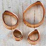 Displayed on a wooden surface, this set of four irregularly shaped metal rings from Kaya Movement—a collection of Teardrop Tunnel Plugs in rose gold—boasts a handcrafted tribal plug design. The 14 gauge teardrop-shaped rings feature smooth edges and a glossy finish that reflects light beautifully.