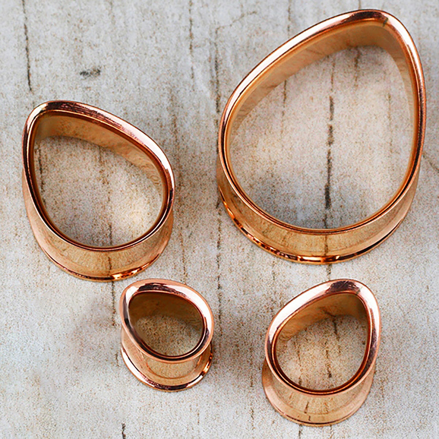 Displayed on a wooden surface, this set of four irregularly shaped metal rings from Kaya Movement—a collection of Teardrop Tunnel Plugs in rose gold—boasts a handcrafted tribal plug design. The 14 gauge teardrop-shaped rings feature smooth edges and a glossy finish that reflects light beautifully.