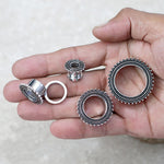 A hand holding several Kaya Movement stainless steel ear gauges in silver and black, featuring intricate beaded designs. The gauges vary in size, with three additional Kaya Movement ear tunnels resting on the palm while one, crafted like a tribal ear tunnel, is held between the thumb and forefinger. The background is a light, neutral surface.