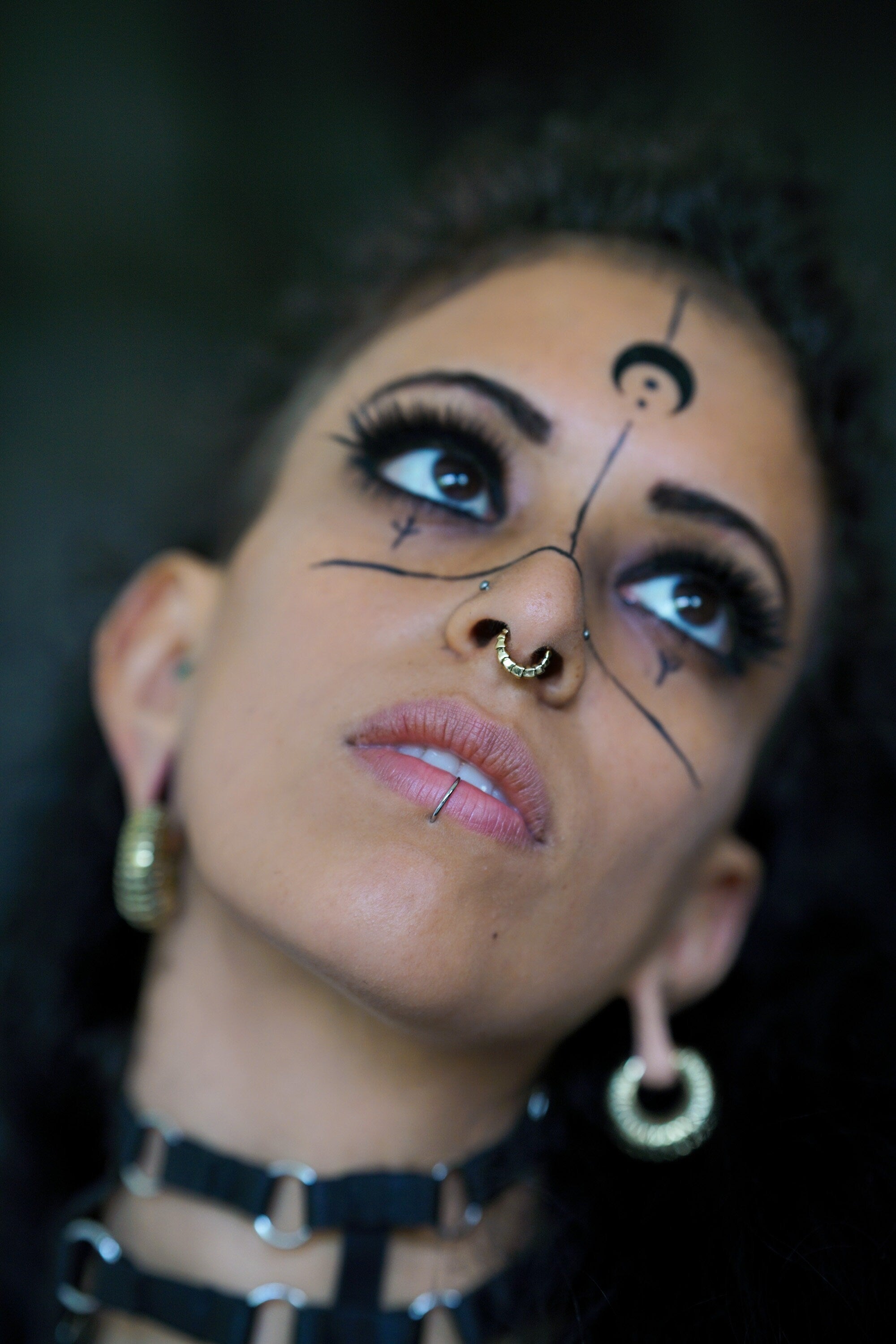 A person with detailed makeup, a Kaya Movement Biomechanical 18k Gold Septum Ring (10mm) featuring an Alien Xenomorph design, and symbolic black markings on their face gazes upwards. They are wearing large hoop earrings and a dark choker necklace with metallic rings. The background is blurred, emphasizing their striking facial features.