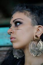 A close-up side profile of a person with intricate face makeup, featuring bold lines and symbols, including a teardrop shape below the eye. They wear large, ornate circular earrings and a Kaya Movement Futuristic Xenomorph 18k Gold-Plated Septum Ring for Men and Women. The background is blurred, with a mix of green and brown hues.