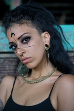 A person with long, dark hair and a partly shaved head gazes into the camera. They have black face paint designs, large hoop earrings, an 18k gold-plated necklace, and a Kaya Movement Futuristic Xenomorph 18k Gold-Plated Septum Ring for Men and Women. They are wearing a black tank top, and the background is blurred with hints of blue and brown.