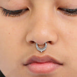 Close-up of a person wearing an ornate silver septum ring. The jewelry, a Silver Septum Ring Clicker Hinged Teardrop 16g by Kaya Movement, features intricate detailing and small decorative accents along its bottom edge. The person has smooth skin and their eyes are gently closed.