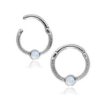 A Kaya Movement Silver Opal Hinged Septum Nose Ring Clicker, designed as an 8mm rook piercing jewelry in 16g stainless steel, features a small round opal gemstone set at the center. The clicker mechanism allows for easy opening and closing. Set against a white background, the reflective surface below casts a subtle shadow, evoking the elegance of fine septum piercings.