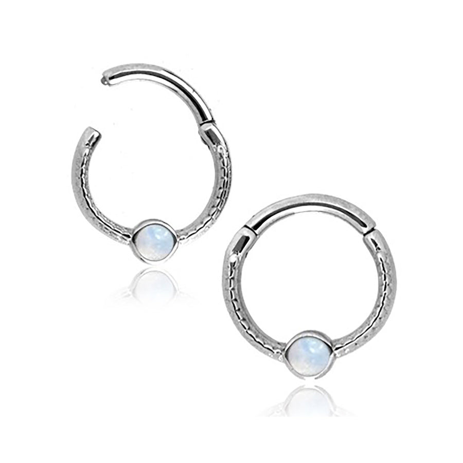 A Kaya Movement Silver Opal Hinged Septum Nose Ring Clicker, designed as an 8mm rook piercing jewelry in 16g stainless steel, features a small round opal gemstone set at the center. The clicker mechanism allows for easy opening and closing. Set against a white background, the reflective surface below casts a subtle shadow, evoking the elegance of fine septum piercings.