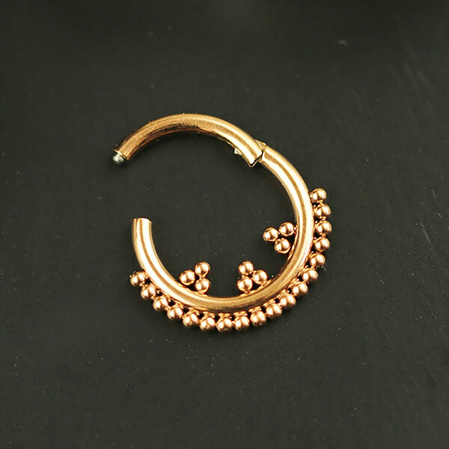 A close-up photograph shows the Rose Gold Septum Clicker 16g - 8mm from Kaya Movement, adorned with small rose gold beads along the bottom half. The septum ring, reminiscent of delicate body jewelry, is set against a dark, smooth background that highlights its intricate detailing and shiny finish.