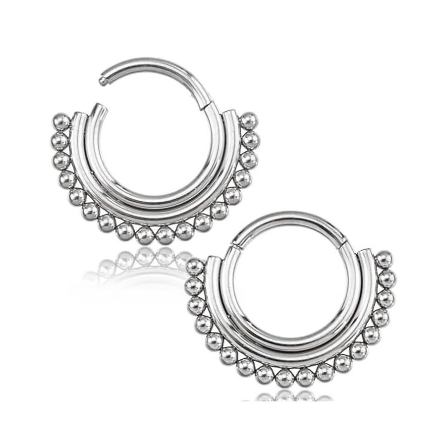 A silver septum clicker, crafted from surgical steel by Kaya Movement, is adorned with small, rounded beads along the outer edge, giving it a decorative and ornate appearance. The hinged design ensures easy wearing. The reflection of the jewelry is visible below it.