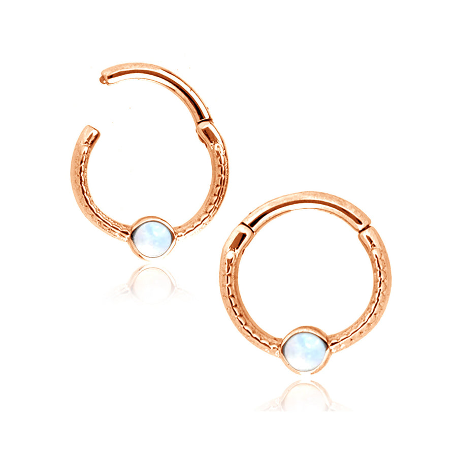 A pair of elegant, circular rose gold earrings, each featuring an opalescent gemstone at the bottom. The earrings have a smooth, polished finish with subtle textured details along the outer edges—reminiscent of the Kaya Movement's Rose Gold Opal Hinged Septum Ring Clicker.