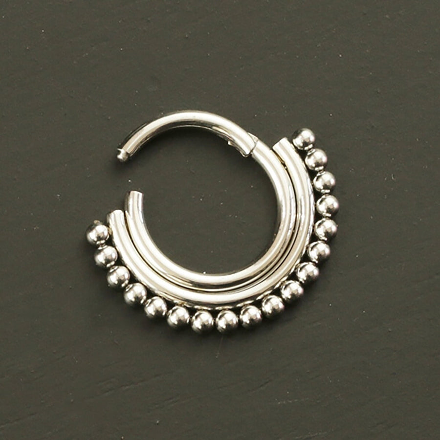The Nose Piercing Surgical steel Septum Clicker 16g - 8mm Cute Silver Hinged Septum Ring from Kaya Movement boasts a bohemian circular design. This surgical steel piece features a series of small silver beads along the outer edge and a smooth, curved bar at the top, forming a half-moon shape. Set against a plain dark background, it pairs perfectly with any septum nose piercing.