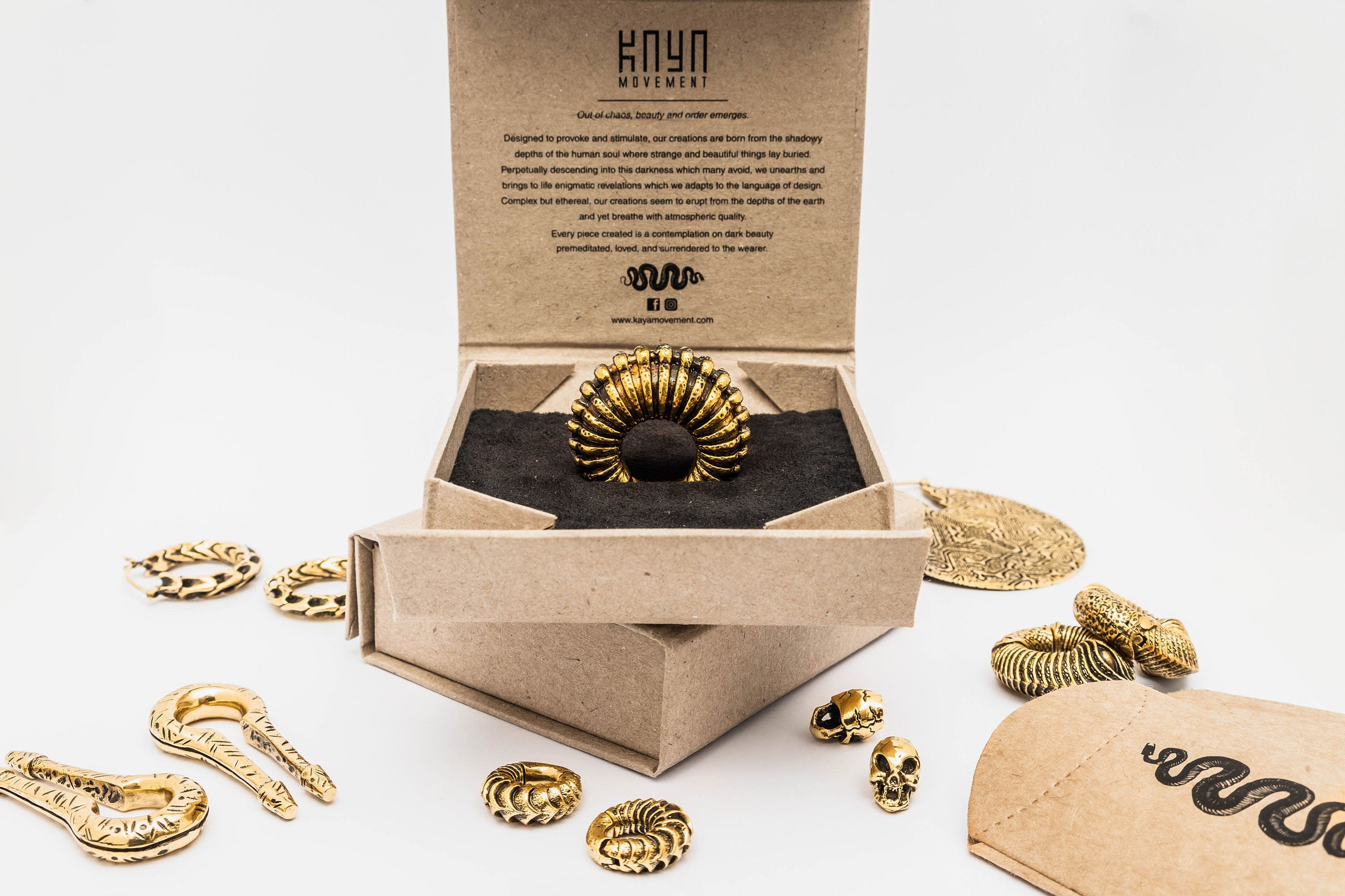 An ornate gold ring is displayed in an open cardboard box with a detailed description inside the lid. Various gold jewelry pieces, including snakes and skull designs, are artistically arranged around the box on a white surface, complemented by intricate body mod jewelry like Kaya Movement's Silver White Brass Ear Weights in keyhole and hanger style.