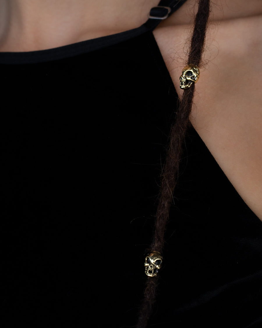 Close-up of a person wearing a black sleeveless top with thin straps. Brown dreadlocks adorned with small, decorative Viking Hair Beads Accessories by Kaya Movement are visible. These Alien Vampire Carved Skull Dreadlocks Beads Jewellery come in a stunning gold finish, adding intricate detail to the hair. The background is blurred, focusing attention on the detailed accessories and fabric texture.
