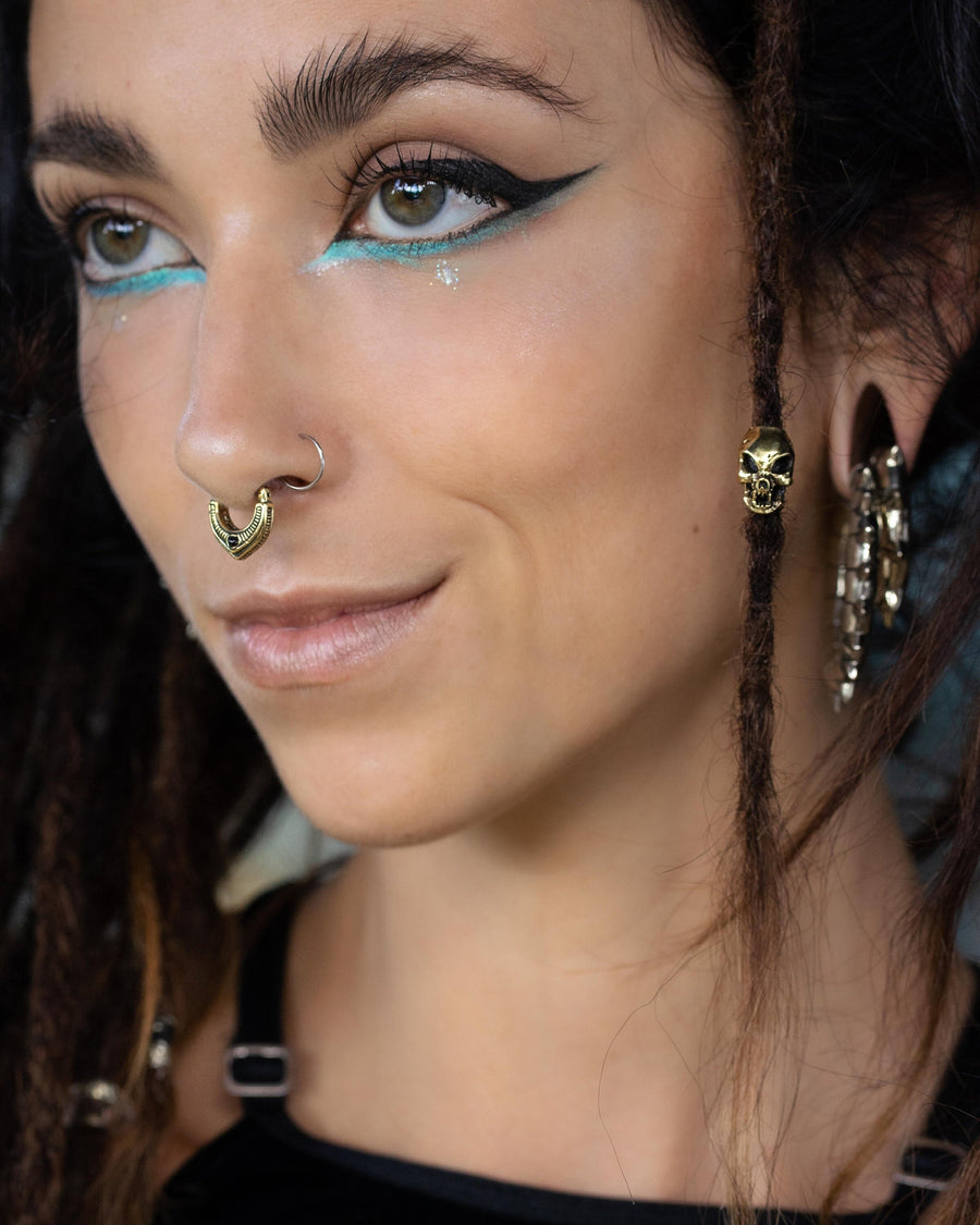 A close-up shot of a woman with brown eyes and bold blue and black eye makeup. She has a nose ring, a septum ring, and is wearing skull-shaped earrings. Her dreadlocks are adorned with brass hair beads and an eye-catching alien vampire carved skull bead from Kaya Movement's Viking Hair Beads Accessories collection, complementing her edgy style in her black top.