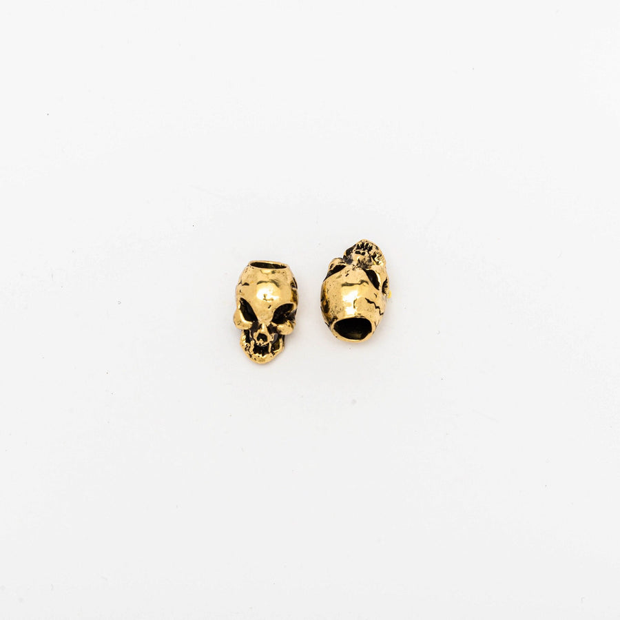 Two small gold-colored charms, perfect as Viking Hair Beads Accessories by Kaya Movement, are placed on a plain white background. These Alien Vampire Carved Skull Dreadlocks Beads Jewellery feature hollowed eyes and nose cavities with detailed facial features. One charm is shown from the front, while the other is displayed at an angle. Ideal for use as dreadlock accessories or unique brass hair beads, these beard accessories add a distinctive touch to your style.