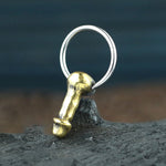 A miniature keychain showcases a gold-hued, metallic charm shaped like an ancient artifact associated with male anatomy, attached to a stainless steel ring. Positioned upright on a textured dark surface against a softly blurred blue background, this unique accessory aligns well with the distinct designs from Kaya Movement's Anatomy Dick Earrings Jewelry Gold collection, which also features hoop earrings and various piercings for men.