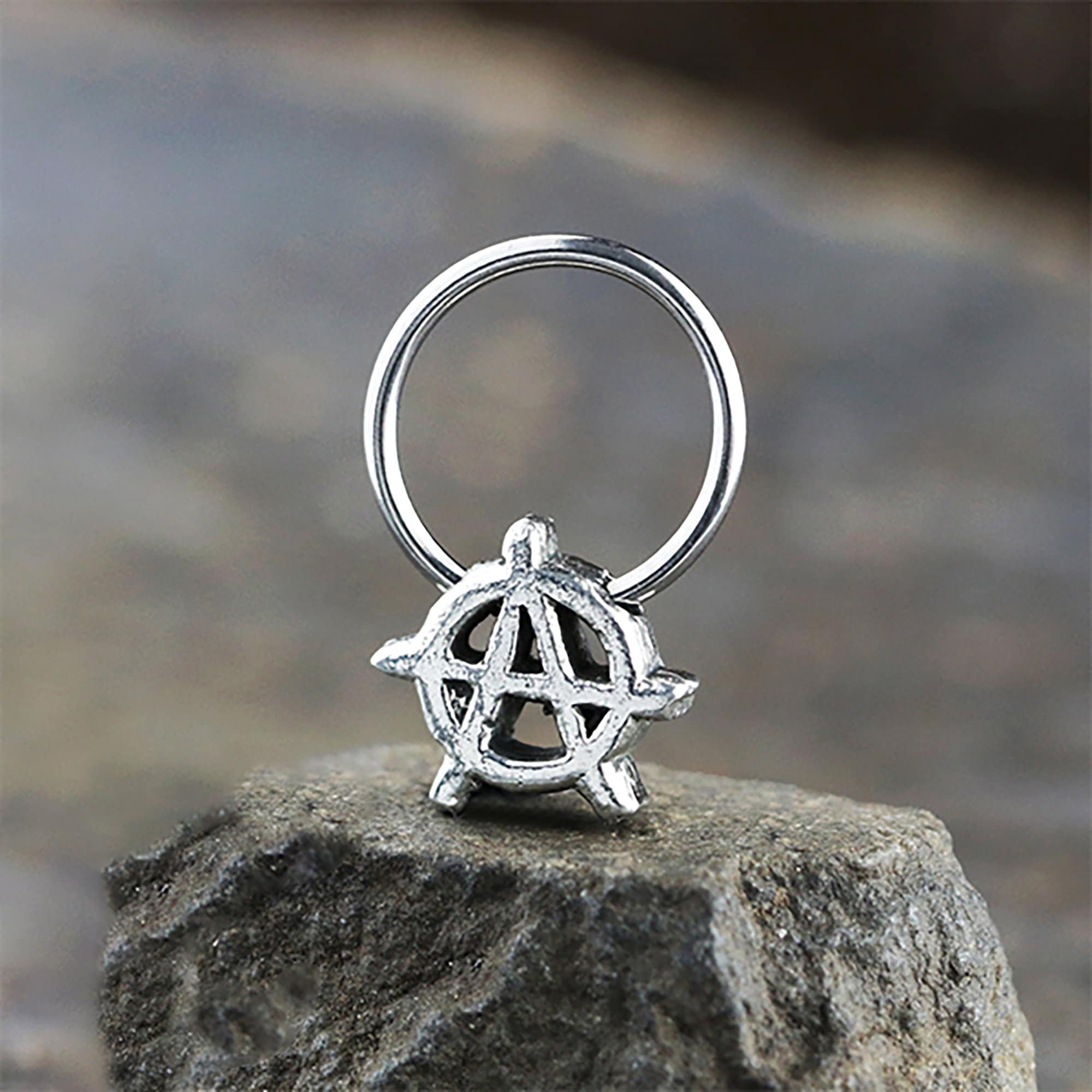 A silver ring with an anarchy symbol on top sits on a rough stone surface. The ring, crafted from Kaya Movement's stainless steel jewelry collection, features a circular motif with an encircled capital "A" at its center, representing the anarchy symbol. The background is blurred, highlighting the texture of the stone and the intricate details of this Anarchy Earrings Jewelry - Silver Septum Ring - Punk Hoop Earrings With Charm Men.