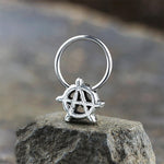 A silver ring with an anarchy symbol on top sits on a rough stone surface. The ring, crafted from Kaya Movement's stainless steel jewelry collection, features a circular motif with an encircled capital "A" at its center, representing the anarchy symbol. The background is blurred, highlighting the texture of the stone and the intricate details of this Anarchy Earrings Jewelry - Silver Septum Ring - Punk Hoop Earrings With Charm Men.