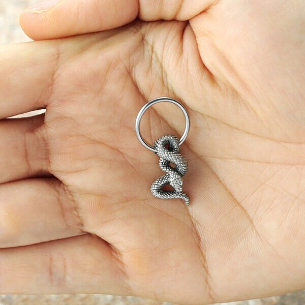 A close-up image of a hand holding a small keychain shaped like an intricately detailed, metallic coiled snake, reminiscent of the exquisite design of Kaya Movement's Silver Snake Hoop Earrings. The keychain loop is attached to the snake's head, echoing the fine craftsmanship seen in septum and helix piercings.