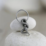 A pair of Silver Snake Hoop Earrings with charm from Kaya Movement gleam against a smooth, off-white stone background. The snake's scales are intricately textured, coiling gracefully around each earring, reminiscent of elegant snake-themed piercing jewelry suitable for helix, daith, and more.