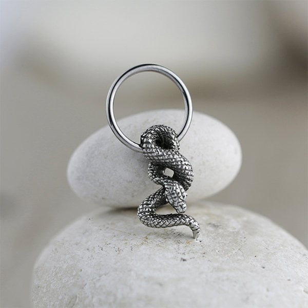 A pair of Silver Snake Hoop Earrings with charm from Kaya Movement gleam against a smooth, off-white stone background. The snake's scales are intricately textured, coiling gracefully around each earring, reminiscent of elegant snake-themed piercing jewelry suitable for helix, daith, and more.