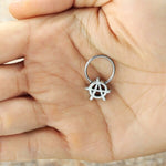 A person's hand is holding a small silver septum ring from Kaya Movement's "Anarchy Earrings Jewelry," made of stainless steel, featuring a pendant shaped like the letter "A" within a circle, resembling the anarchist symbol. The background is blurred, emphasizing the hand and the ring.
