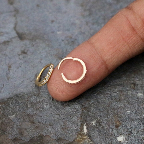 A person holds a small Kaya Movement Hinged Septum Ring Clicker (16g) adorned with tiny cubic zirconia stones, shaped like a hoop, with their index finger. Another identical earring lies on a textured gray surface. These versatile earrings are perfect for nose piercings and various other body piercings such as conch, daith, helix, cartilage, and rook.