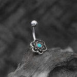 The Turquoise Belly Ring from Kaya Movement is a stunning Silver 925 curved barbell featuring a lotus flower design on one end and a ball at the other. The flower showcases intricate details with black accents and a striking turquoise center, making it perfect for your navel piercing. This exquisite piece of body jewelry stands out beautifully against a textured, dark background.