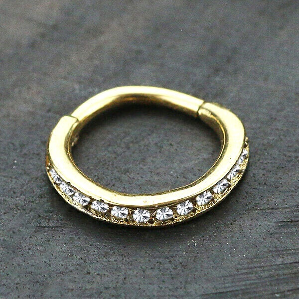A small gold Hinged Septum Ring Clicker by Kaya Movement, adorned with sparkling cubic zirconia gemstones across the front, is placed on a dark, textured surface. Its design is simple yet elegant, ideal for nose piercings.