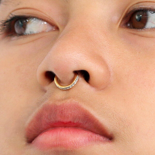 Close-up of a person's face showcasing the Kaya Movement Hinged Septum Ring Clicker 16g. The gold septum ring is adorned with cubic zirconia on one side, enhancing its elegance. The person has light skin, slightly parted lips, and eyes that gaze to the side.
