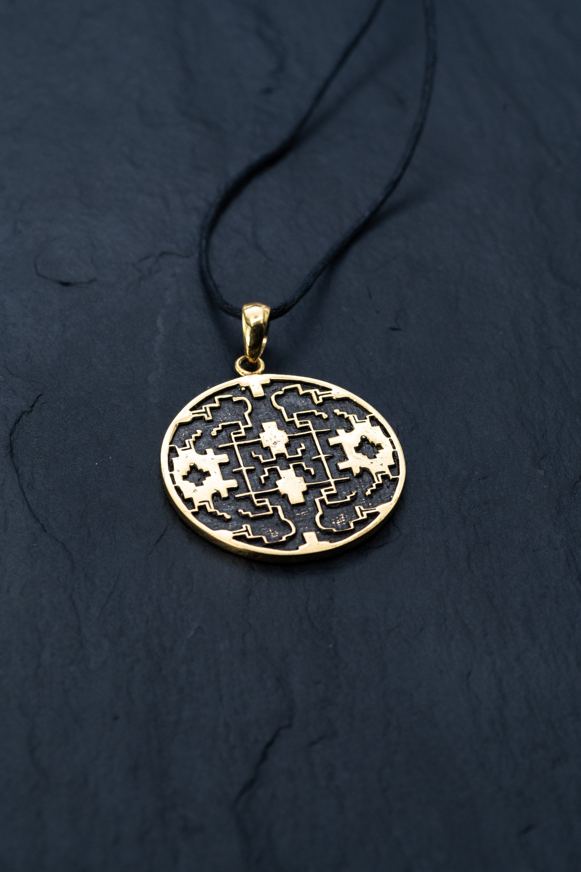 The Shipibo pendant from Kaya Movement, crafted from yellow brass, showcases intricate, maze-like designs and oxidized details. This striking gold pendant hangs elegantly from a black cord against a dark, textured background, featuring symmetrical patterns and geometric shapes that create an eye-catching visual effect.