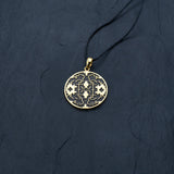 A round Shipibo pendant from Kaya Movement's Shamanic jewelry collection rests on a thin black cord against a dark textured background. This intricate Icaros pendant showcases a detailed geometric design in oxidized yellow brass and black, featuring symmetrical patterns with small cross shapes and other decorative elements.
