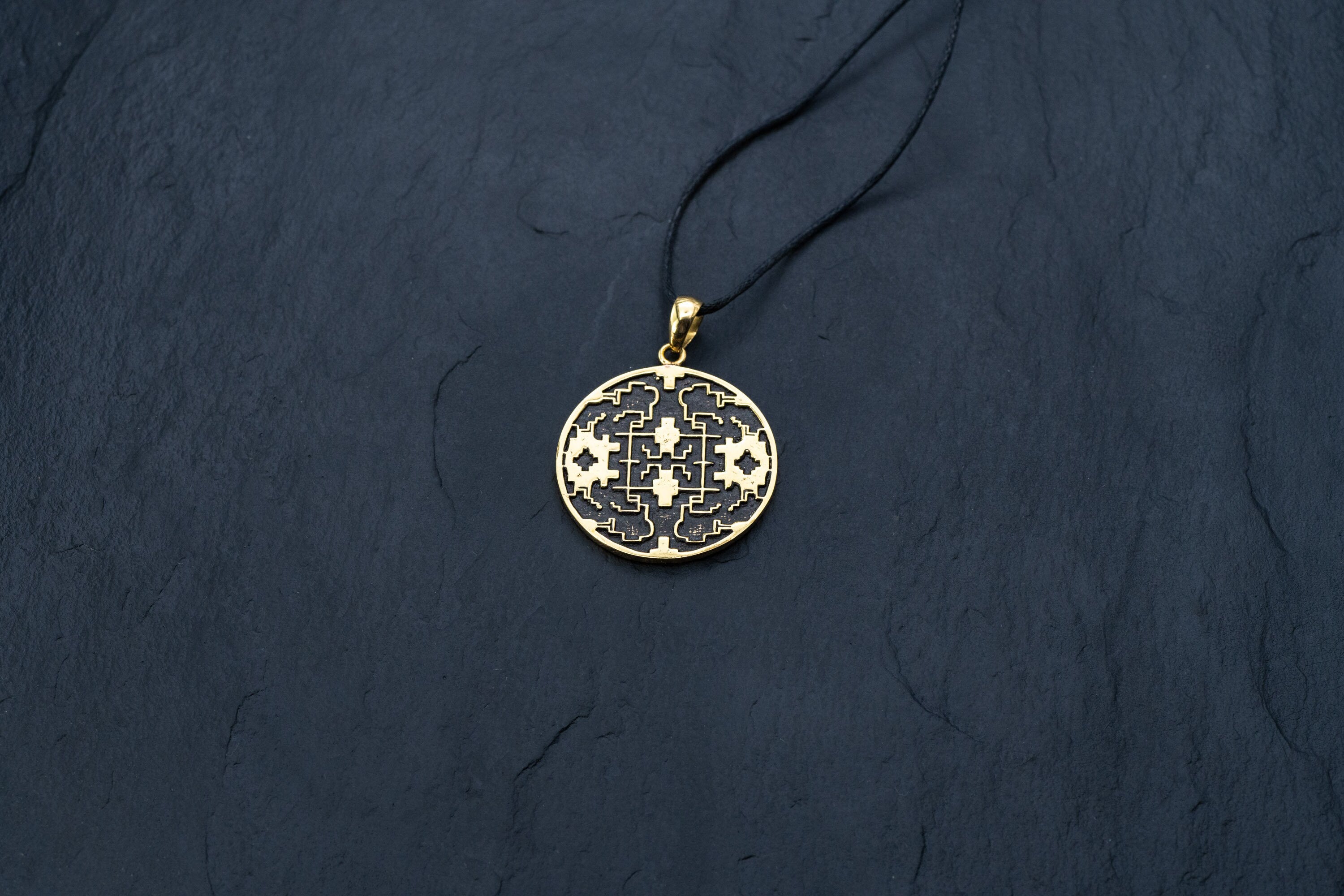 A round Shipibo pendant from Kaya Movement's Shamanic jewelry collection rests on a thin black cord against a dark textured background. This intricate Icaros pendant showcases a detailed geometric design in oxidized yellow brass and black, featuring symmetrical patterns with small cross shapes and other decorative elements.